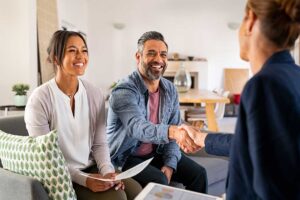 A real estate agent discussing property details with a couple at a table filled with papers and a rural house view through the window.